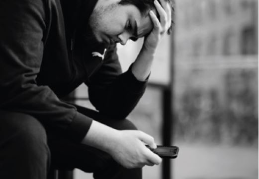 En person som sitter utomhus på en bänk med mobiltelefonen i handen och ser nedstämd ut